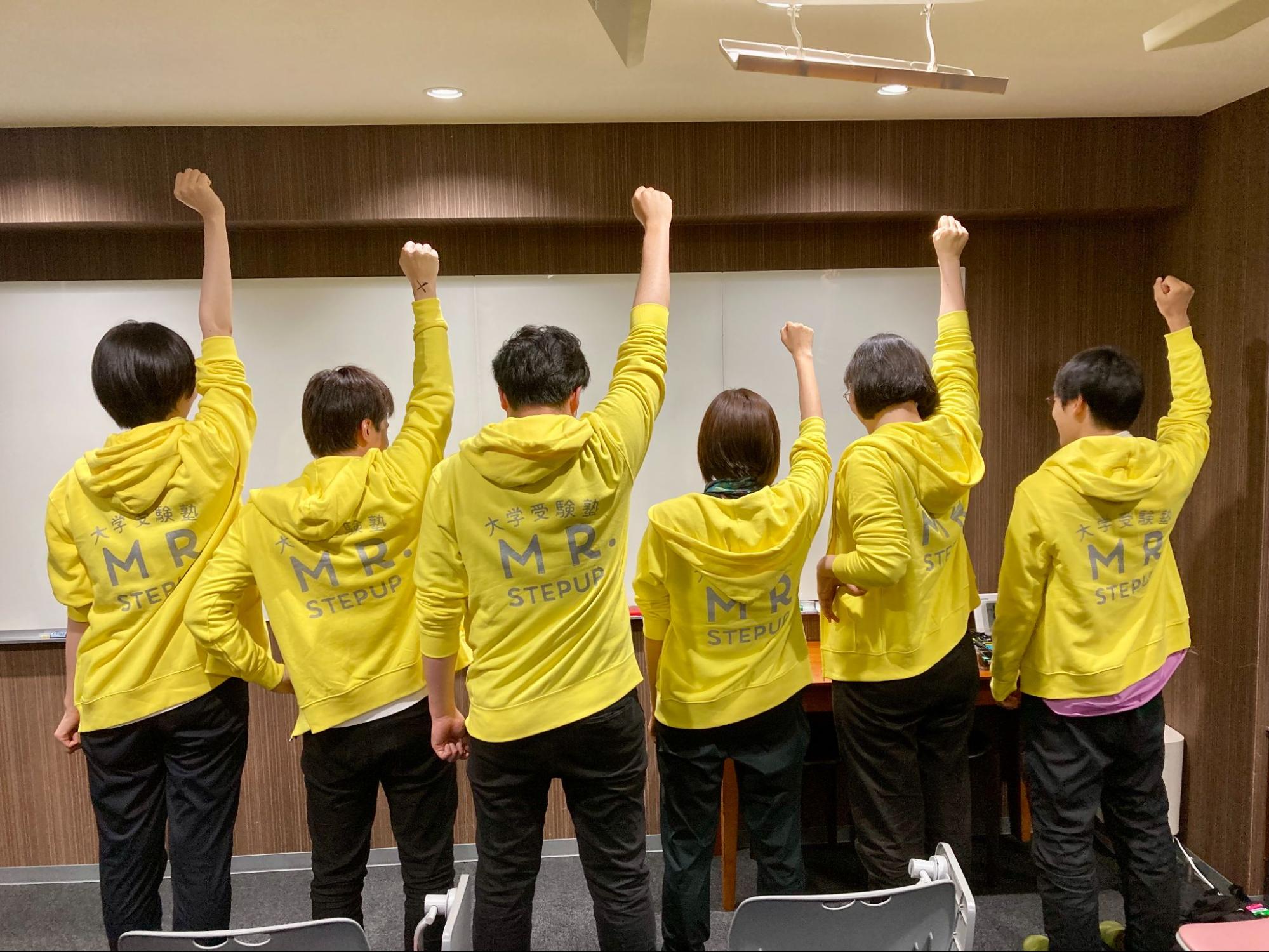 塾のスタッフたちが黄色いパーカーを着て後ろを向き、ONE PIECEのシーンのように腕を上げている集合写真