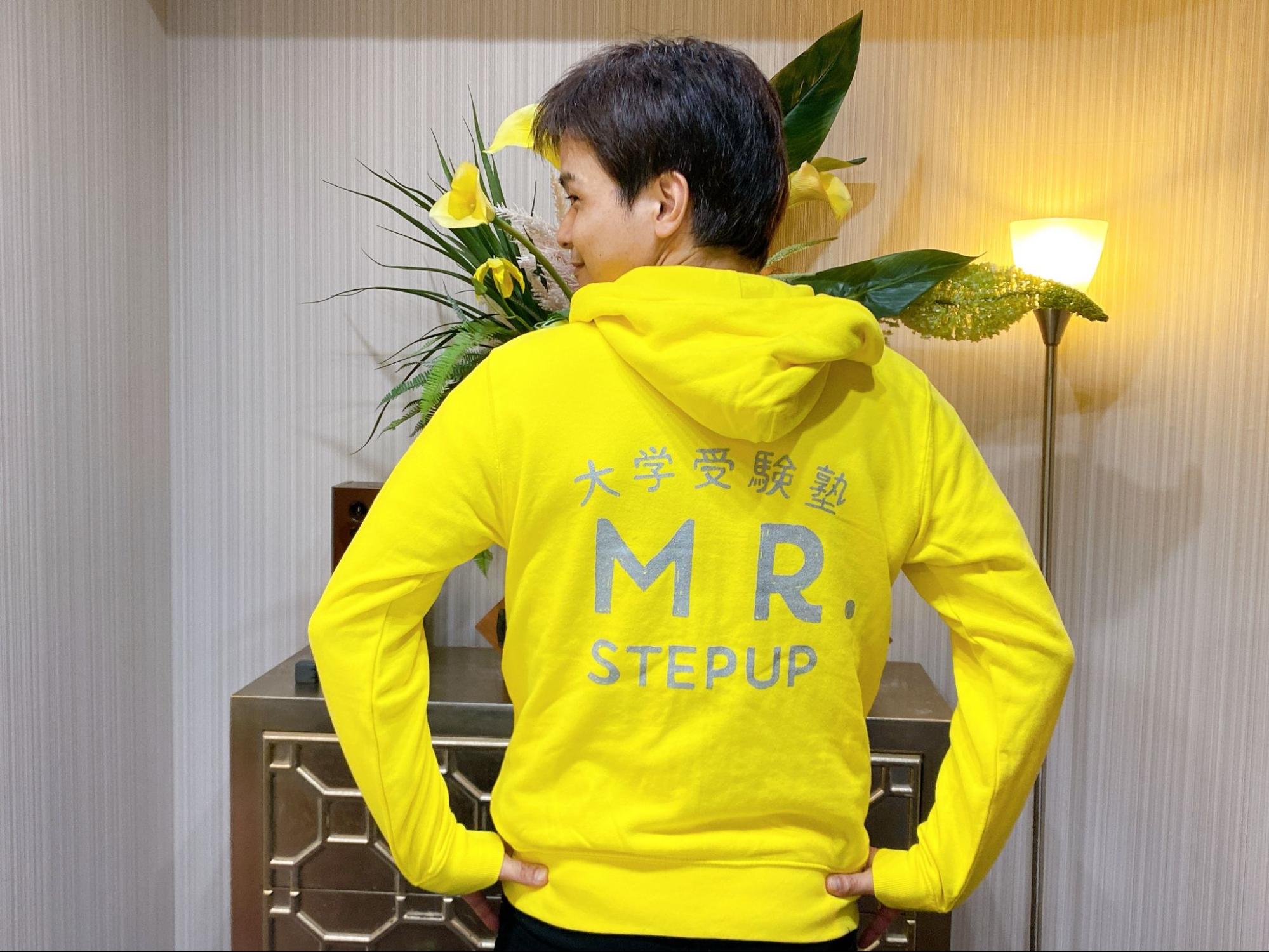 講師よなたんが黄色いパーカーを着て後ろを向いている写真、背中には「大学受験塾 MR. STEPUP」の文字